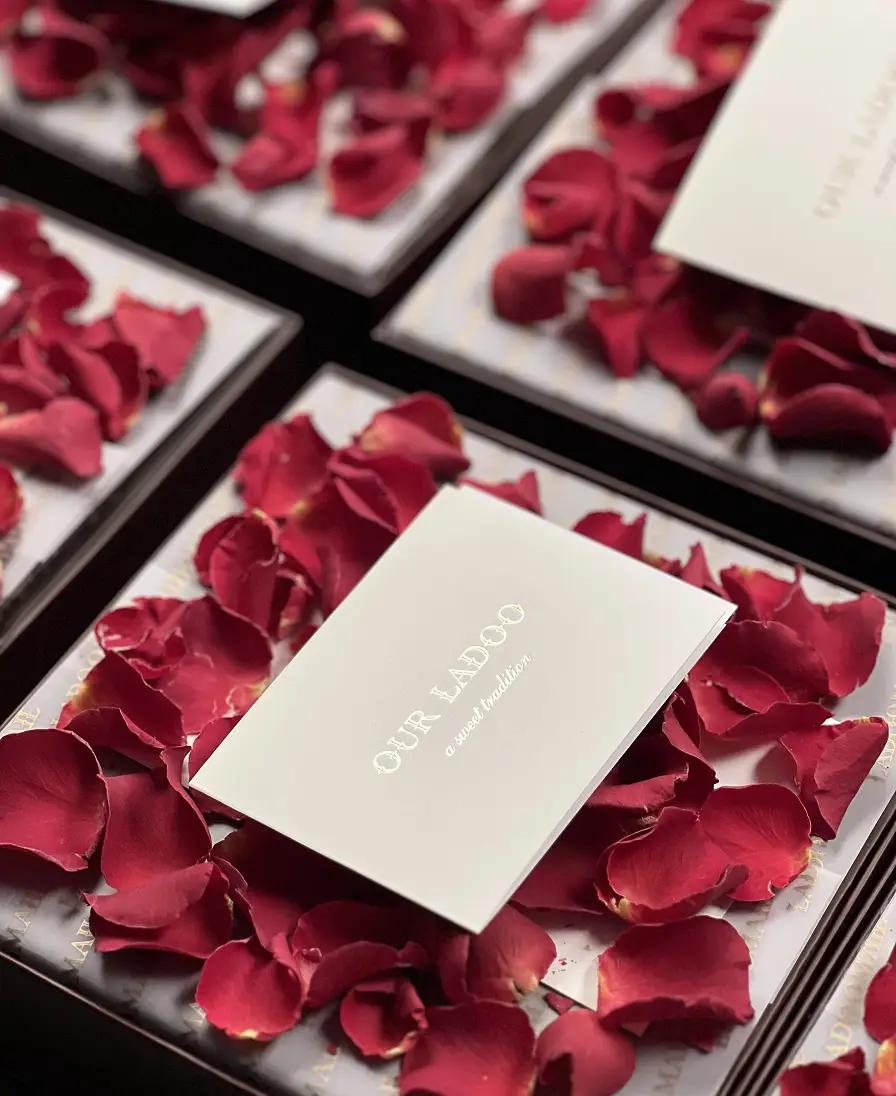 The Gift Box Ladoomail burgundy gift boxes open with rose petals and 'Our Ladoo, a sweet tradition' cards.