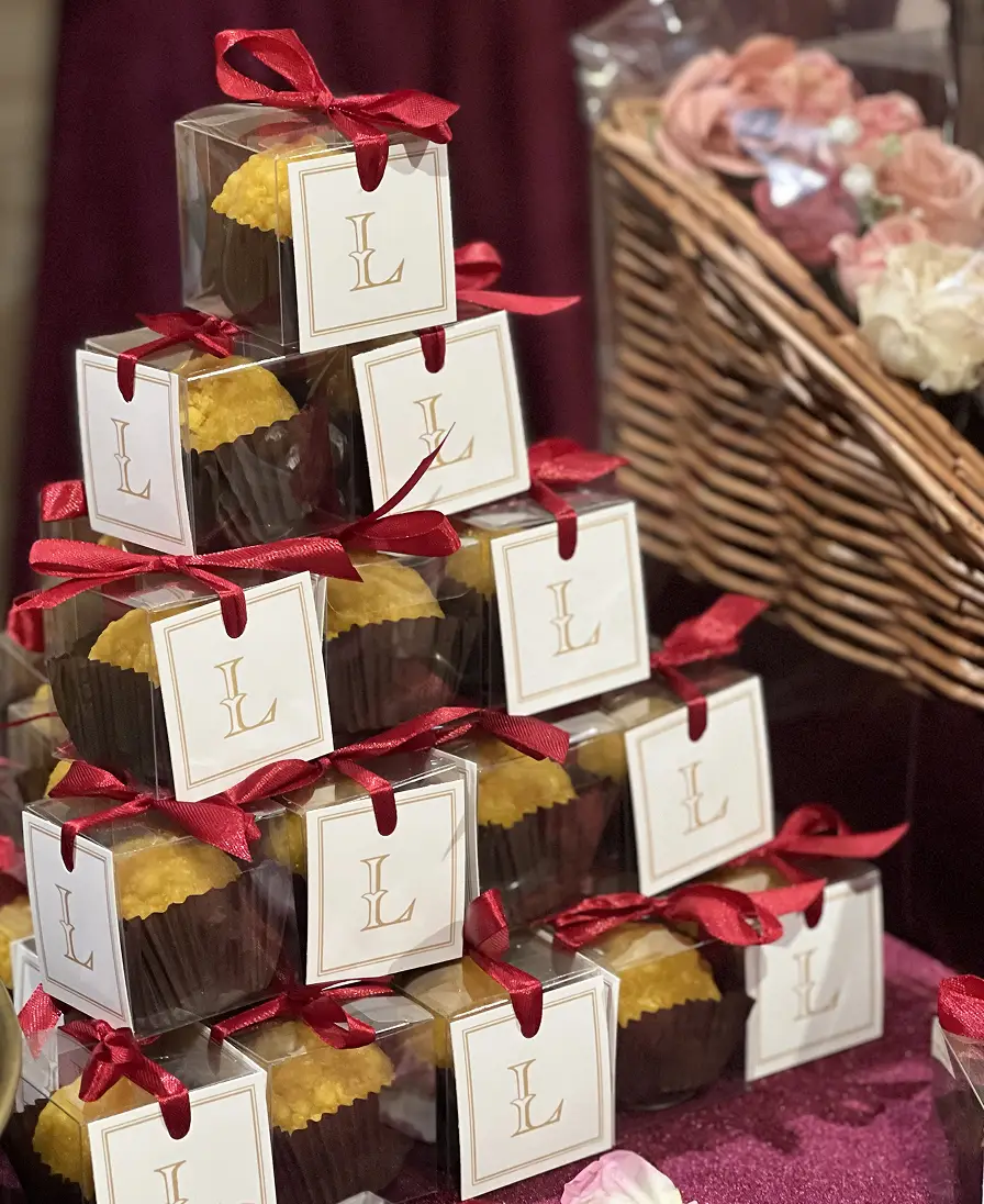 Celebration favours Stacked Ladoomail celebration favours with monogrammed tags and red ribbon beside a flower-filled wicker basket.